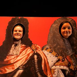 Two people smile with their faces showing through cutouts in a painted board depicting figures in elaborate royal costumes, reminiscent of those worn during the Battle of Aughrim, set against a vivid red background.