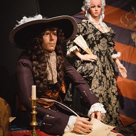 Two mannequins in elaborate 17th-century attire—one seated at a desk with a quill and paper, the other standing behind in a patterned gown—set against a backdrop featuring a historic Battle of Aughrim flag.