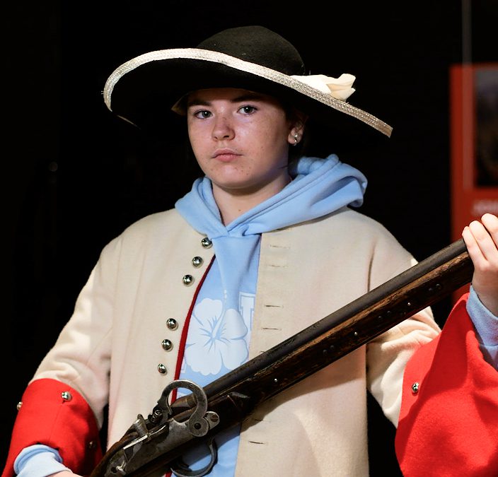 A young person wearing a historical military-style coat, a wide-brimmed hat with a ribbon, and a blue hoodie holds an old-fashioned musket, evoking the Battle of Aughrim as they look seriously at the camera against a dark background.