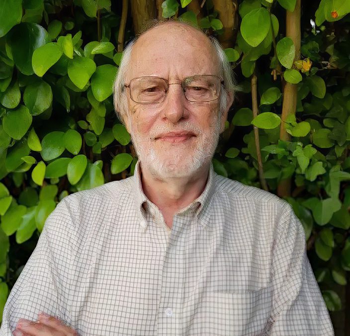 An older man with glasses and a white beard stands in front of lush green foliage, wearing a light plaid shirt and smiling gently with his arms crossed—his calm demeanor reminiscent of a historian reflecting on the Battle of Aughrim.