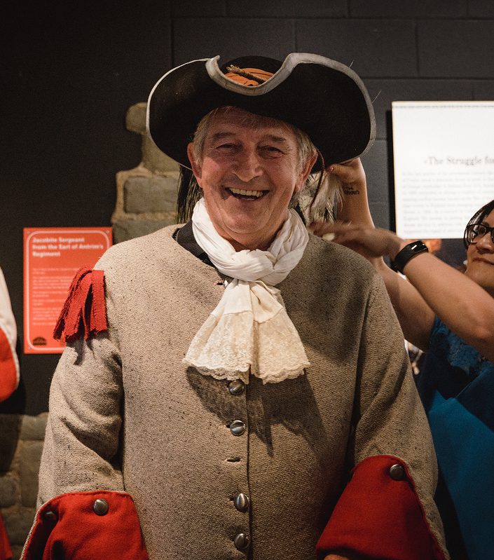 An older man smiles while wearing a historical military costume with a tricorne hat, beige coat with red cuffs, and a white neck scarf—evoking the era of the Battle of Aughrim. People stand behind him, adjusting his outfit.