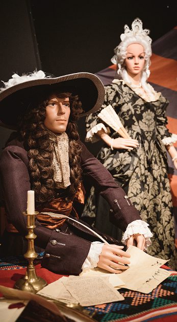 Two mannequins in historical attire evoke the era of the Battle of Aughrim: one seated at a desk writing with a quill, wearing a curly wig and wide-brimmed hat; the other stands behind in an ornate dress and lace headdress, holding a fan.