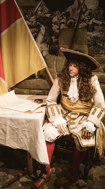 A man in elaborate 17th-century clothing with a large hat and curly wig sits at a table draped with a white cloth, next to a yellow and red flag, in a historical setting suggestive of the Battle of Aughrim, with a mural behind him.