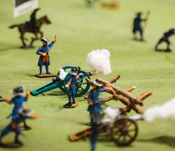 Small toy soldiers in blue uniforms are arranged on a green surface, recreating the Battle of Aughrim with cannons and cotton for smoke. A horse and more figures add depth to this historic scene.