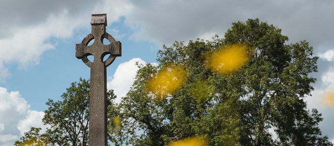 A stone Celtic cross stands outdoors against a backdrop of green trees and a cloudy sky, with yellow-blurred flowers in the foreground—evoking memories of the Battle of Aughrim.