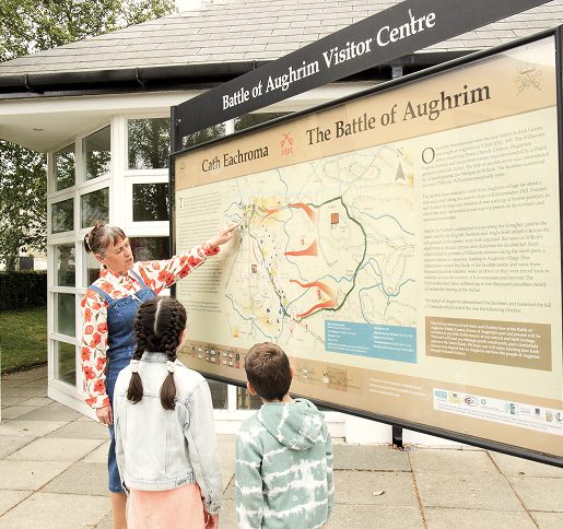 A woman points at a large Battle of Aughrim map display outside the Visitor Centre, while two children stand in front of her, listening intently and studying the historic battlefield layout.