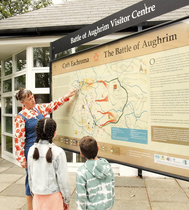 A woman points to a large Battle of Aughrim informational map at an outdoor visitor center, while two children stand in front of her, listening attentively.