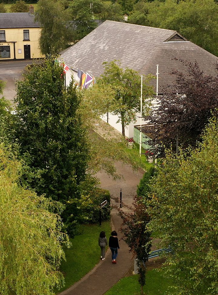 Two people walk along a winding path through a green park with trees. Nearby, a building with a gray roof and several flags, including the Union Jack, flies—hinting at the area’s connection to the Battle of Aughrim. Another building is in the background.
