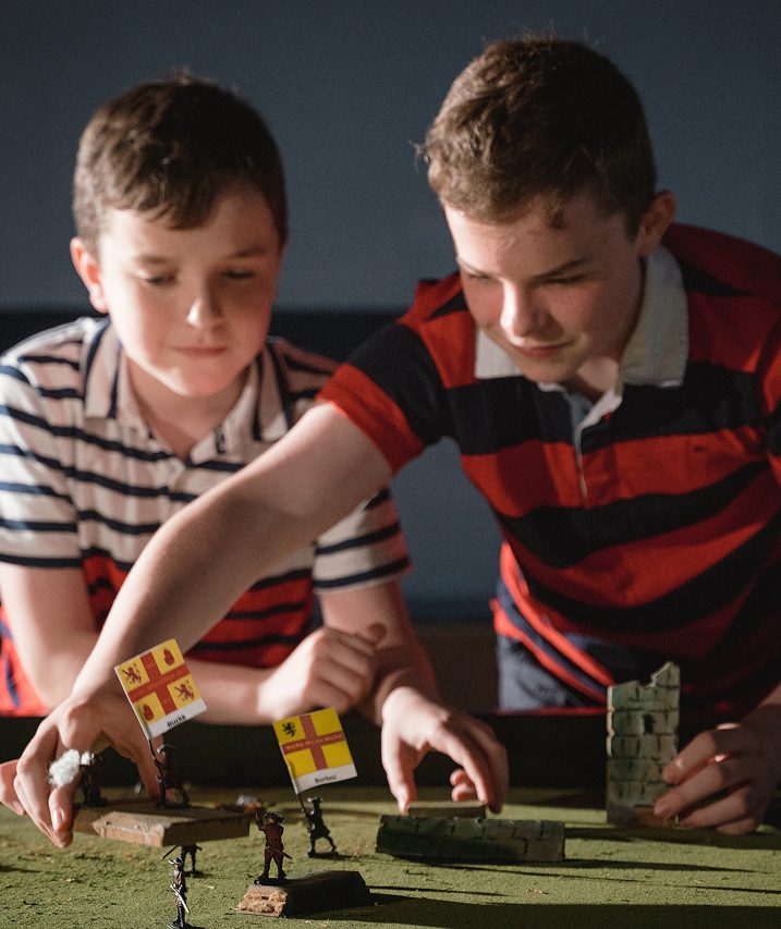 Two boys in striped shirts play a tabletop war game, moving miniature soldiers and flags across a green surface with small model walls, re-enacting the Battle of Aughrim as they focus intently on their game.