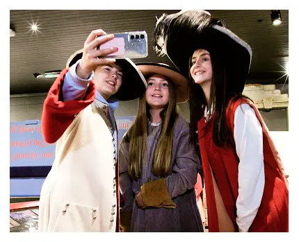 Three smiling teens dressed in historical costumes and large hats take a selfie together inside a museum, with informational signs and displays about the Battle of Aughrim in the background.