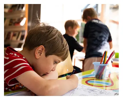 A young boy in a red striped shirt is drawing at a table with colored pencils, while two other children stand and interact in the background, discussing the Battle of Aughrim in a brightly lit classroom.