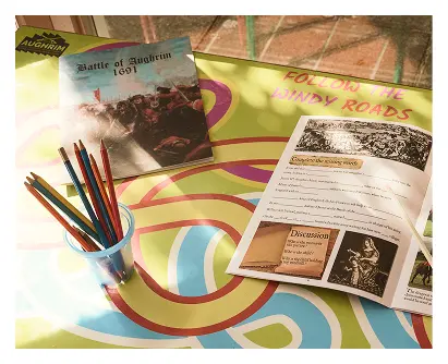 A colorful table displays an open booklet, the book Battle of Aughrim 1691, and a cup of colored pencils. FOLLOW THE WINDY ROADS is printed on the table as sunlight streams in, evoking echoes of the historic Battle of Aughrim.